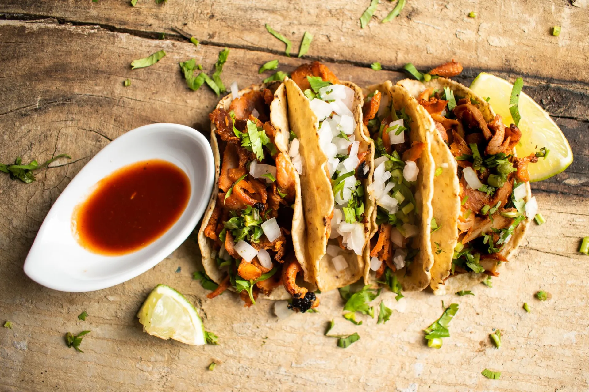 Four street tacos on corn tortillas, topped with onion and cilantro, served with a lime wedge and a basket of tortilla chips.